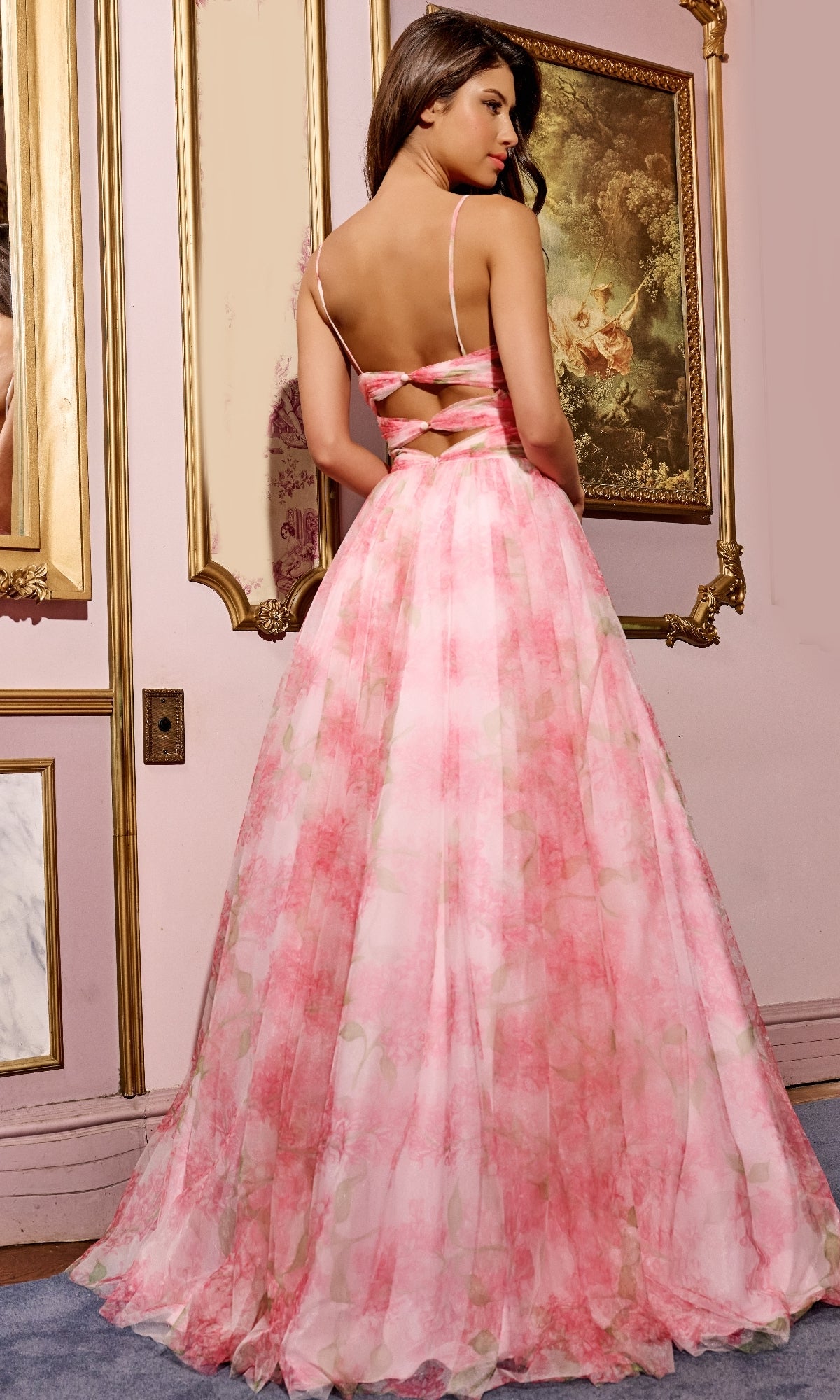 A woman in a pink Cut-Out Long Floral-Print Prom Dress (Jovani 40402) with thin straps stands in a vintage-style room decorated with ornate gold-framed paintings.